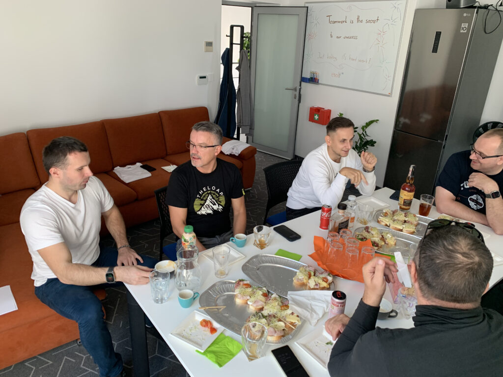 A casual office gathering with team members sitting around a table covered in drinks and open-faced sandwiches, having a relaxed conversation; a whiteboard with a humorous team slogan is visible in the background.