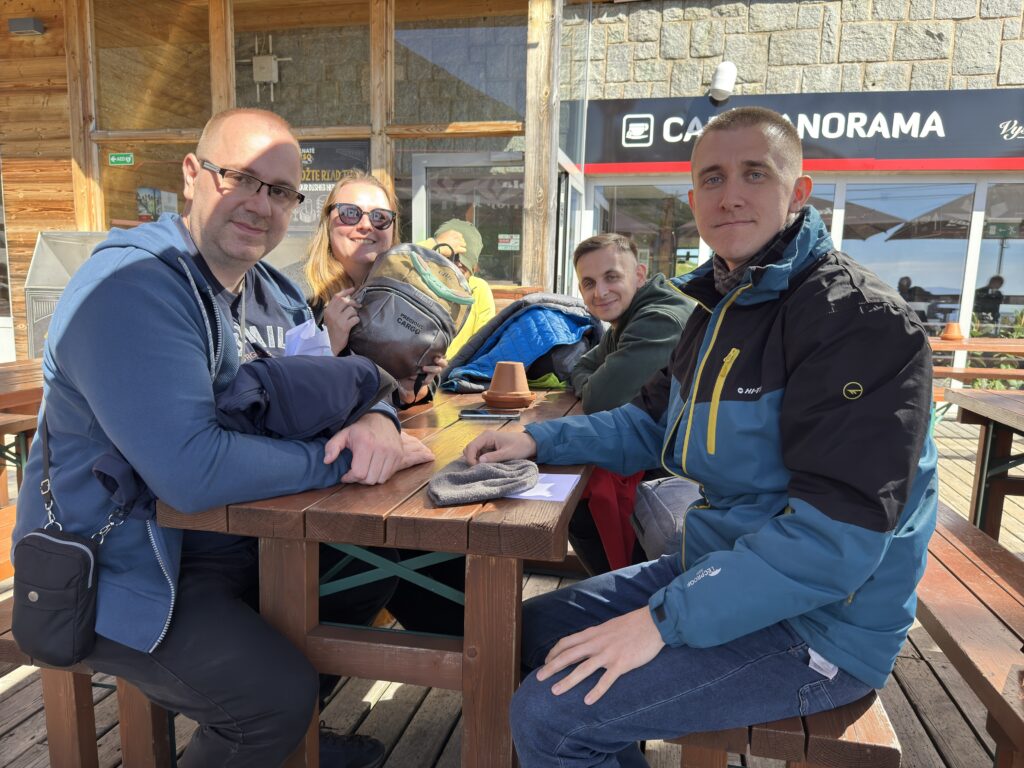 Group of five people sitting at a wooden table outside Café Panorama in the mountains, smiling and enjoying a sunny day