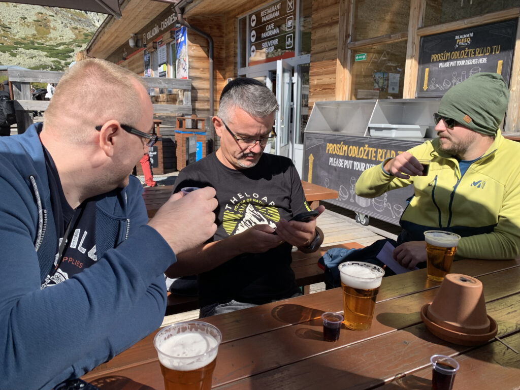 Three men sitting at an outdoor table in front of a mountain café, drinking beer and shots, with one man focused on his phone