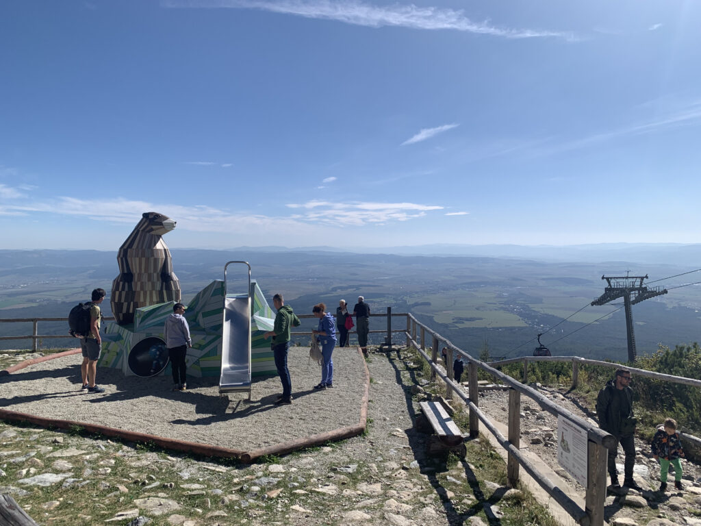 People standing near a children's slide shaped like an animal sculpture on a mountain viewpoint, with a cable car tower and wide valley view in the background