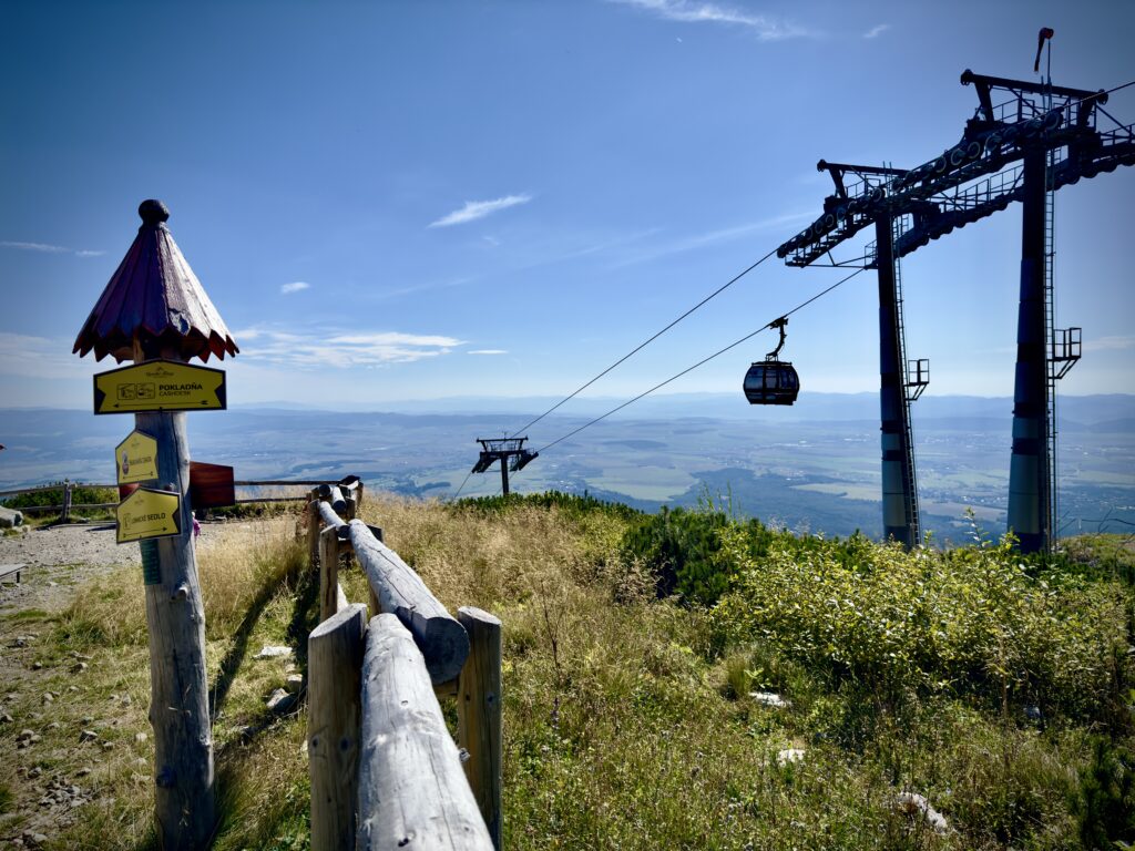 Cable car ascending above a green mountain slope with directional signs and wooden fence in the foreground, and a wide valley view in the background