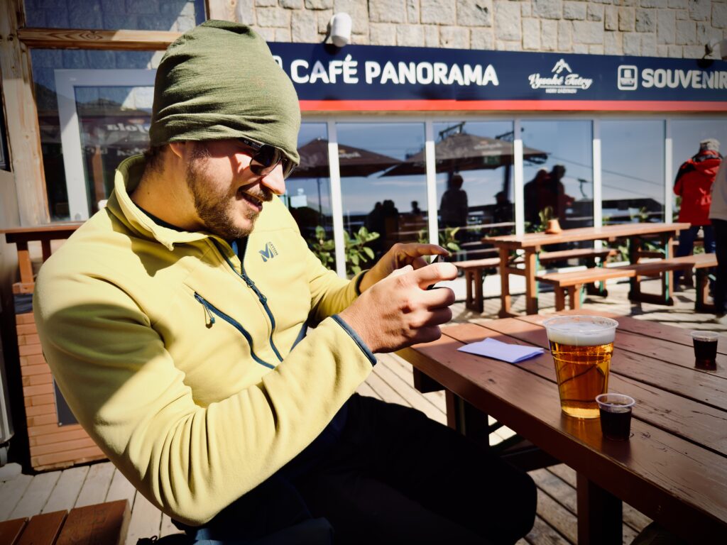 Man in a yellow jacket and green beanie sitting at an outdoor table, smiling while looking at his phone, with a beer and shots in front of him at Café Panorama in the High Tatras