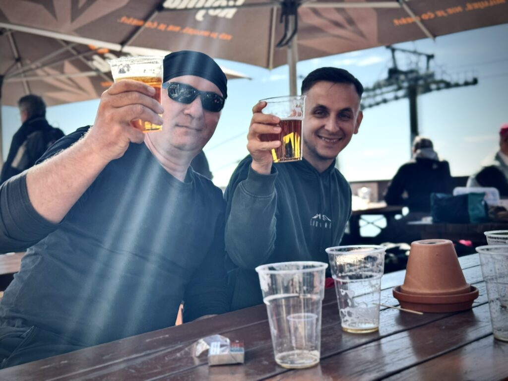 Two smiling men toasting with beer at a wooden outdoor table, with cable car infrastructure and sunbeams in the background