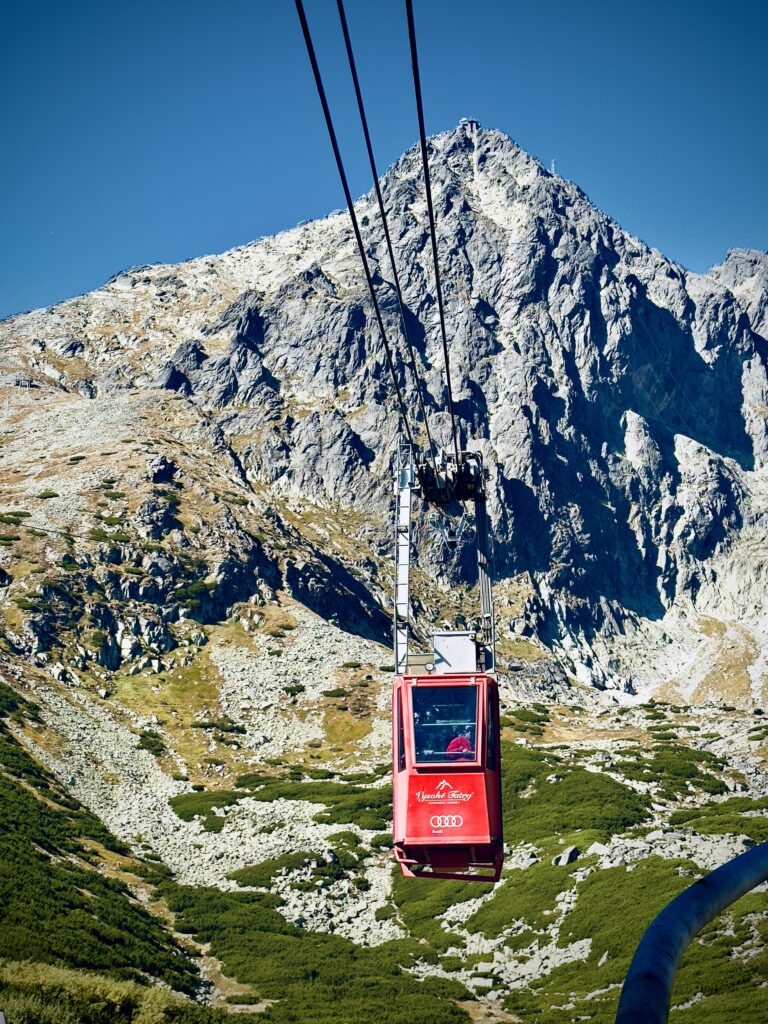 Red cable car suspended mid-air on its way up a rocky mountain slope in the High Tatras, with Lomnický štít peak visible in the background