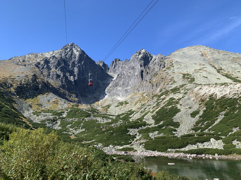 Red cable car ascending toward Lomnický štít peak, surrounded by rocky slopes and green patches above a mountain lake in the High Tatras