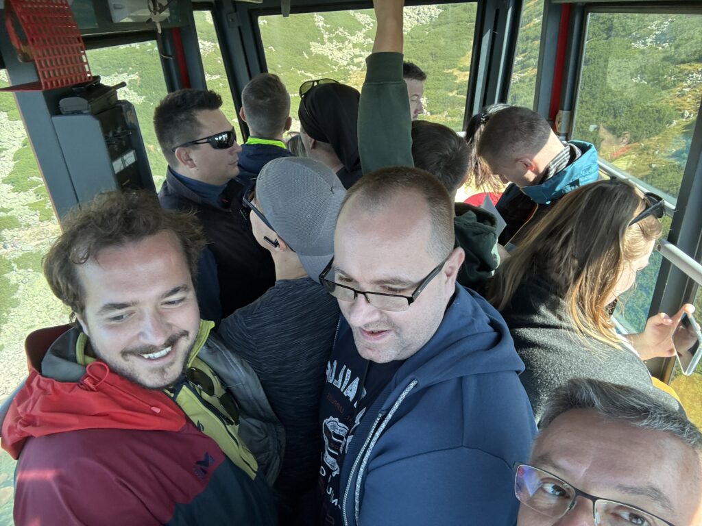 Group of people tightly packed inside a cable car, smiling and enjoying the ride up a mountain with green hills visible through the windows