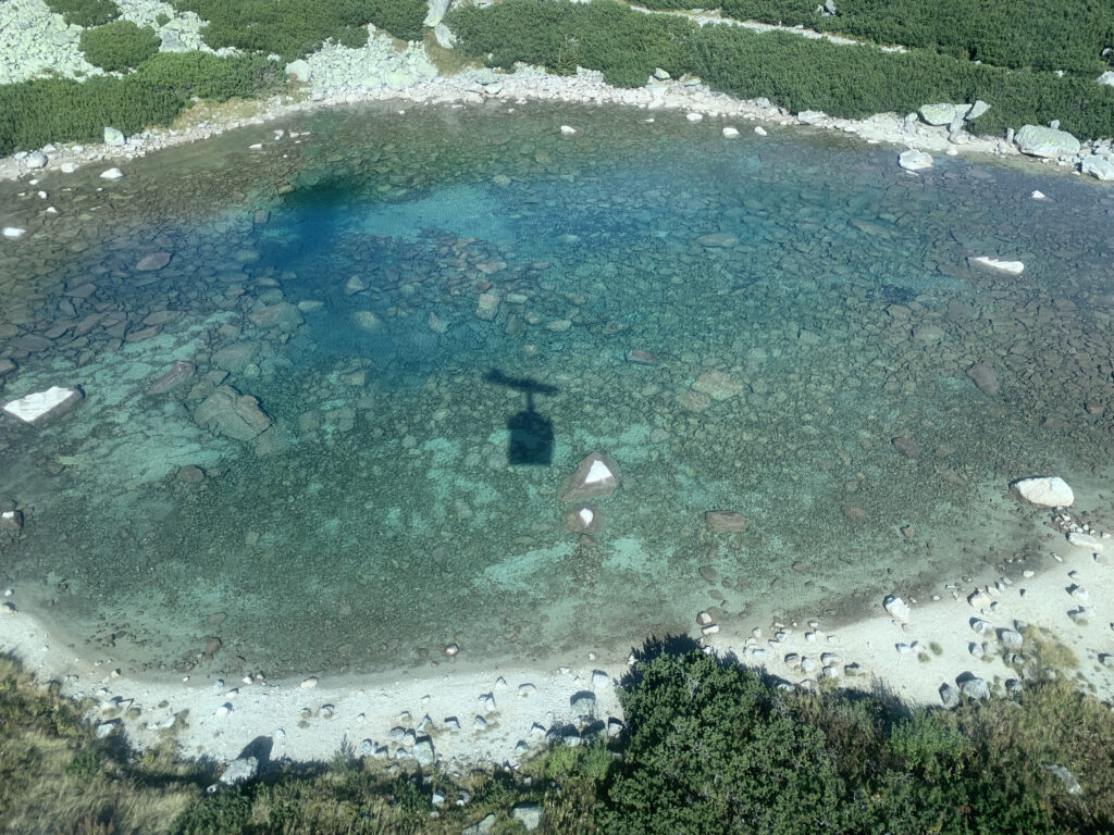 Crystal-clear alpine lake with rocks visible under the surface and the shadow of a cable car cast on the water