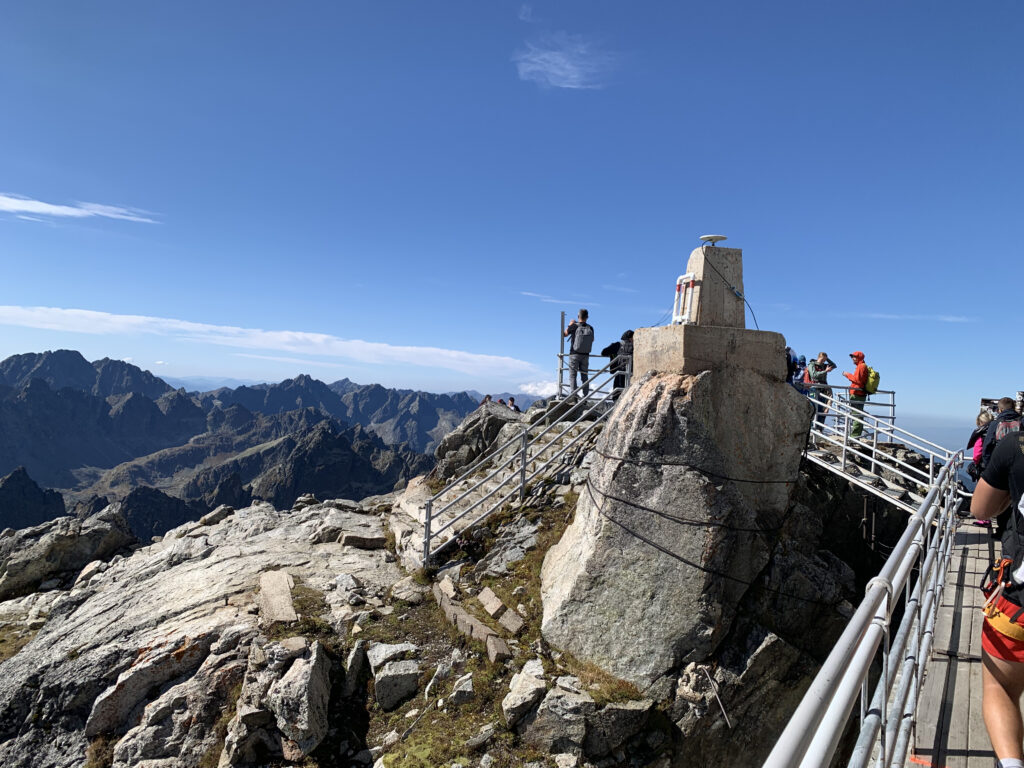 Hikers standing near a summit marker surrounded by railings on a rocky mountain peak with panoramic views of the High Tatras