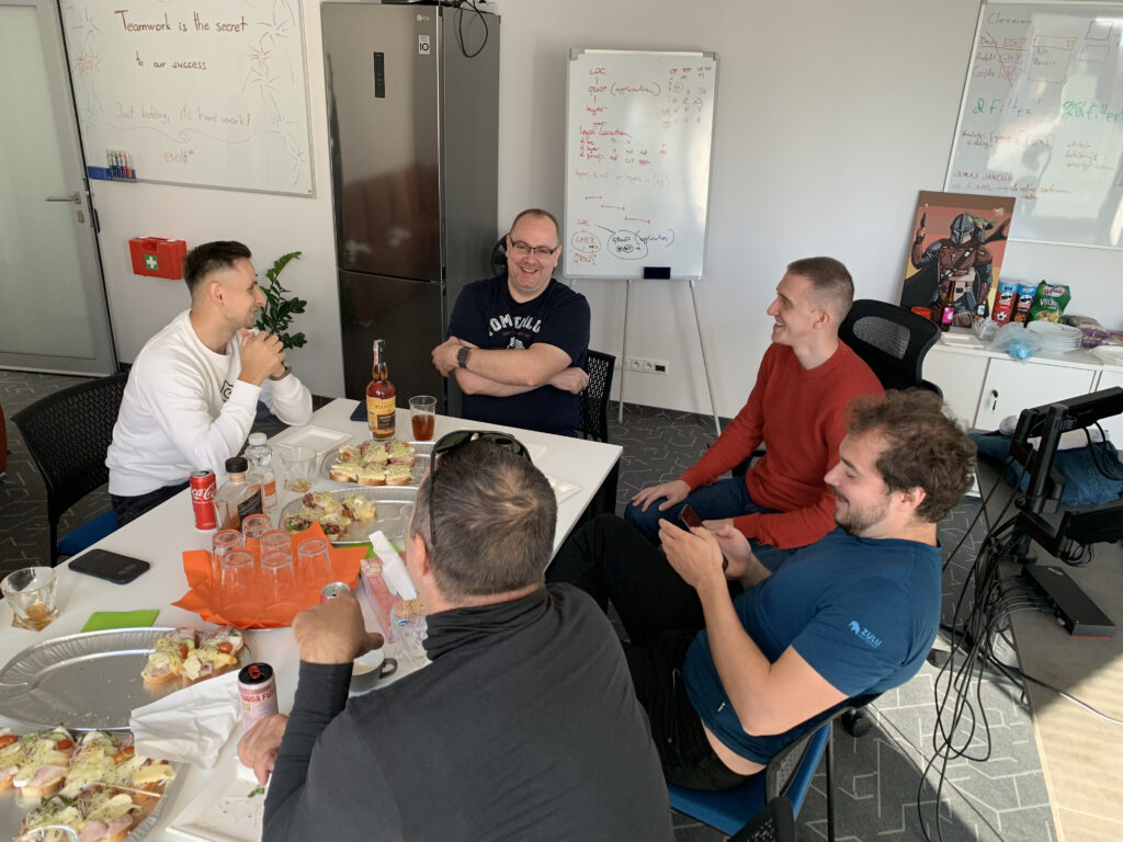A group of colleagues gathered around a table in an office, sharing food and drinks while laughing and chatting; whiteboards with notes and a motivational sign are visible in the background.