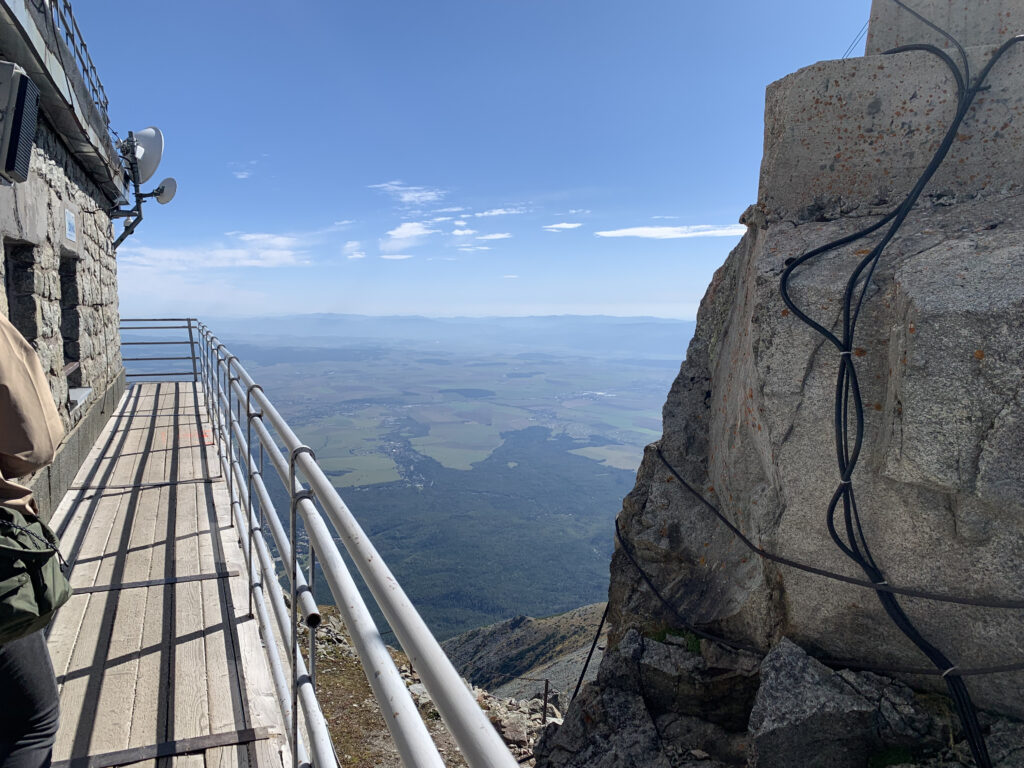 Narrow walkway with railing alongside a stone observatory building on a mountain peak, overlooking a vast valley and farmlands