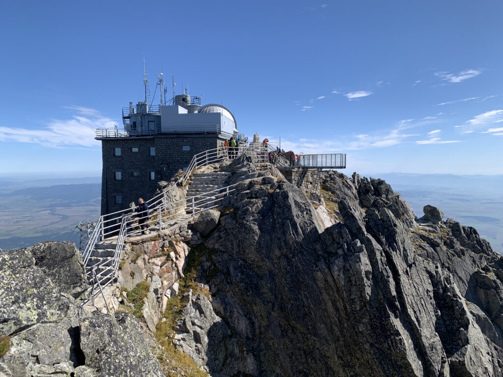 Stone observatory building with antennas and railings perched on a rugged mountain peak, with people walking along the path under a clear sky