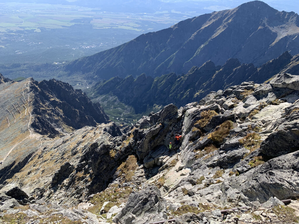 Mountain slope with a zigzagging hiking trail and two hikers in bright jackets climbing among rocks, with a wide valley and forest in the background