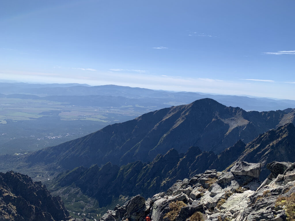 Mountain view with layered ridges, a wide valley with fields and forests, and a hazy blue horizon under a clear sky