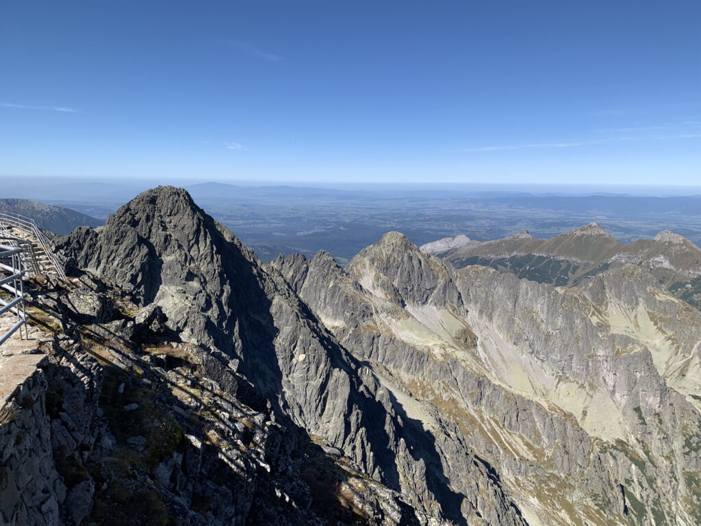 View from a mountain observation point with a railing, overlooking jagged rocky peaks and a vast valley under a clear sky