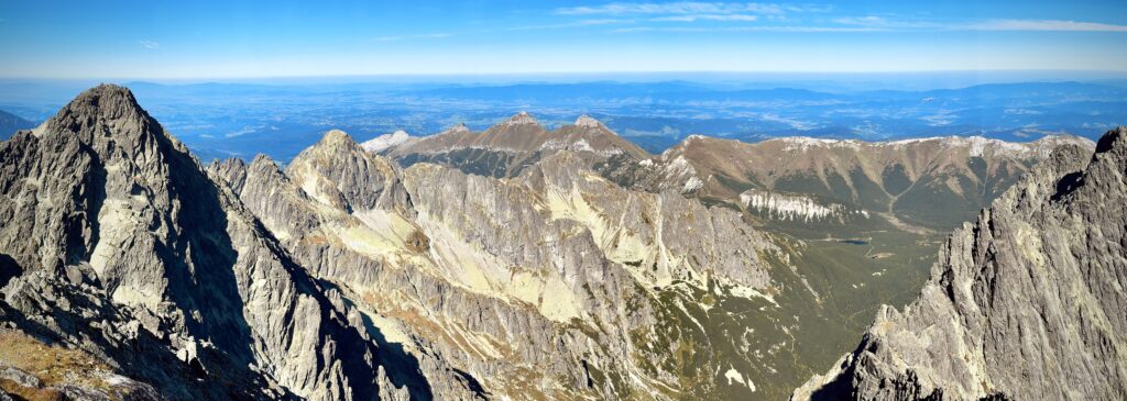 Panoramic view of the High Tatras mountain range with sharp rocky peaks, grassy ridges, and distant valleys under a clear blue sky
