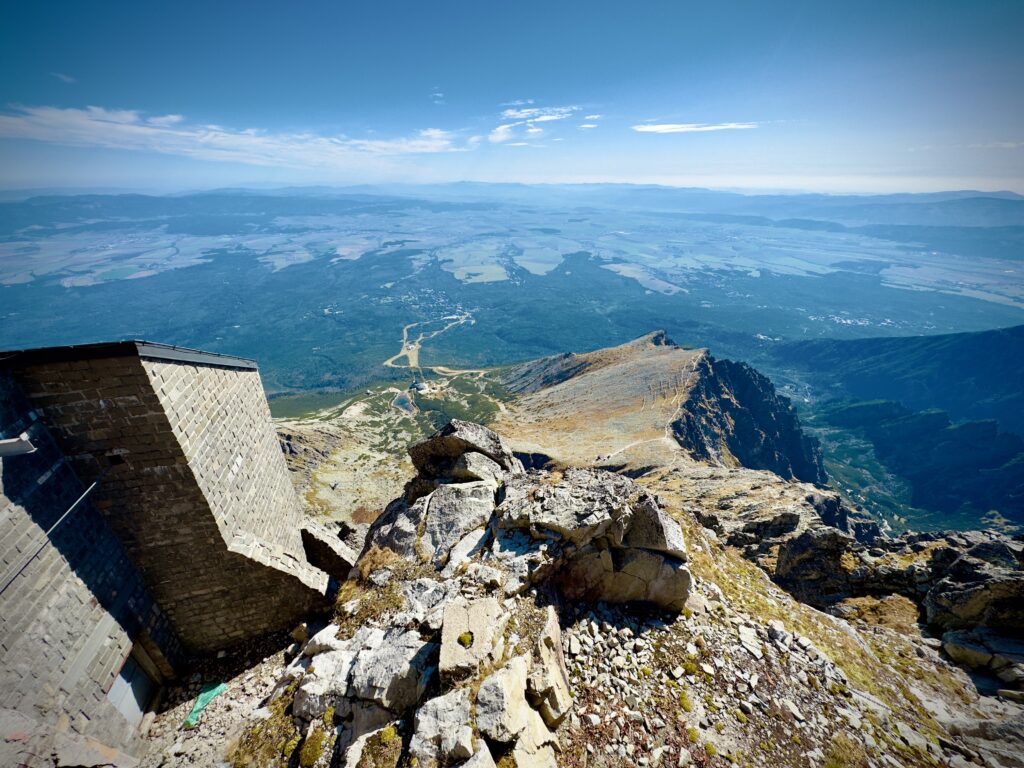 Three men standing at a mountain viewpoint behind a railing, looking out over a wide valley and distant hills under a clear blue sky