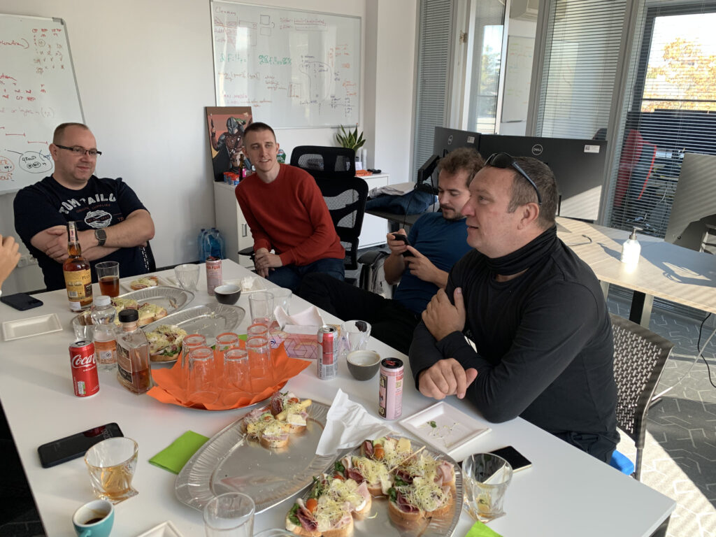 A group of men sitting around a table in an office, enjoying open-faced sandwiches, drinks, and conversation; whiteboards with diagrams are visible in the background.