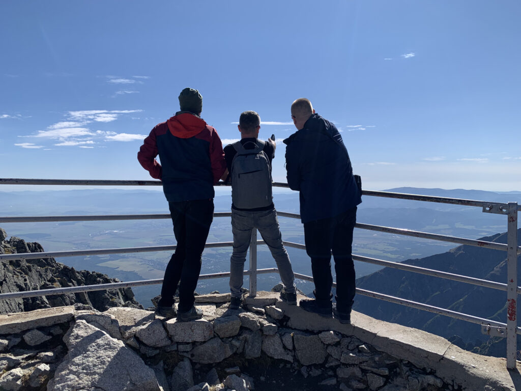 Three men standing at a mountain viewpoint behind a railing, looking out over a wide valley and distant hills under a clear blue sky