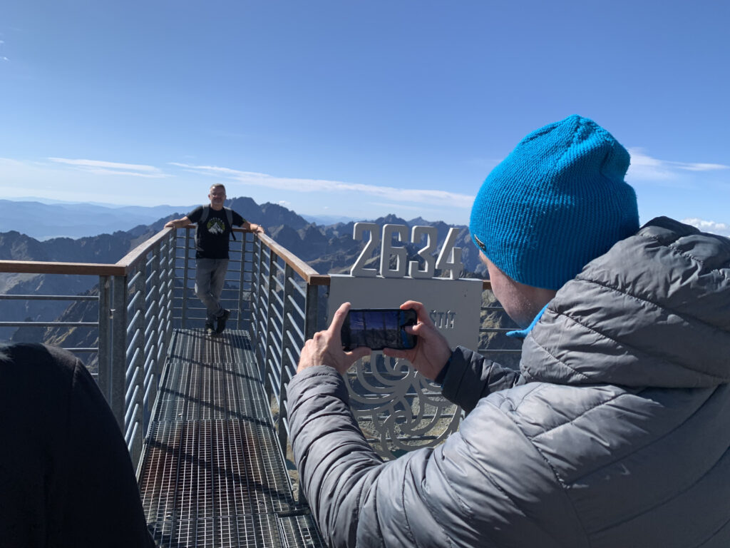 Man taking a photo of a colleague posing on the Lomnický štít viewing platform at 2634 meters with mountain peaks in the background