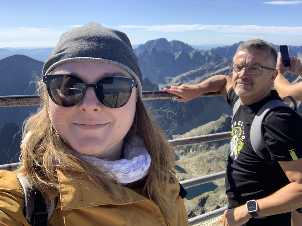 Two people taking a selfie at a mountain viewpoint with rocky peaks, blue sky, and a mountain lake visible in the background