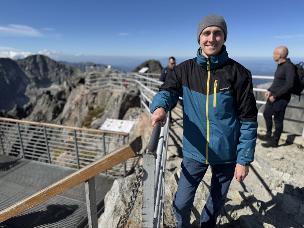 Man in a blue jacket and gray beanie smiling at a mountain viewpoint with dramatic rocky peaks and safety railings around
