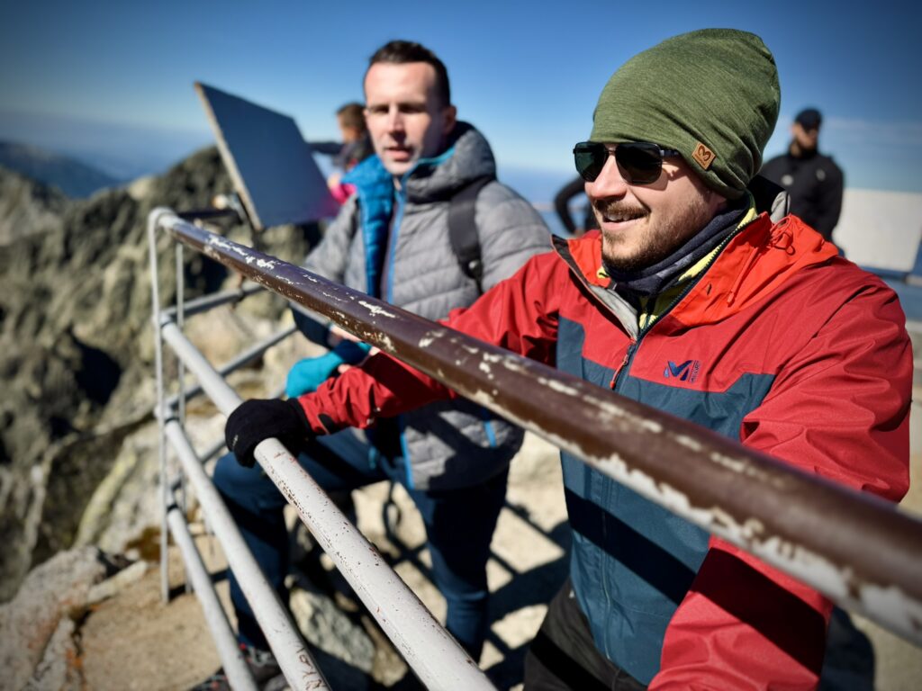 Two men in outdoor gear standing at a mountain viewpoint behind a metal railing, enjoying the sunny weather and panoramic views