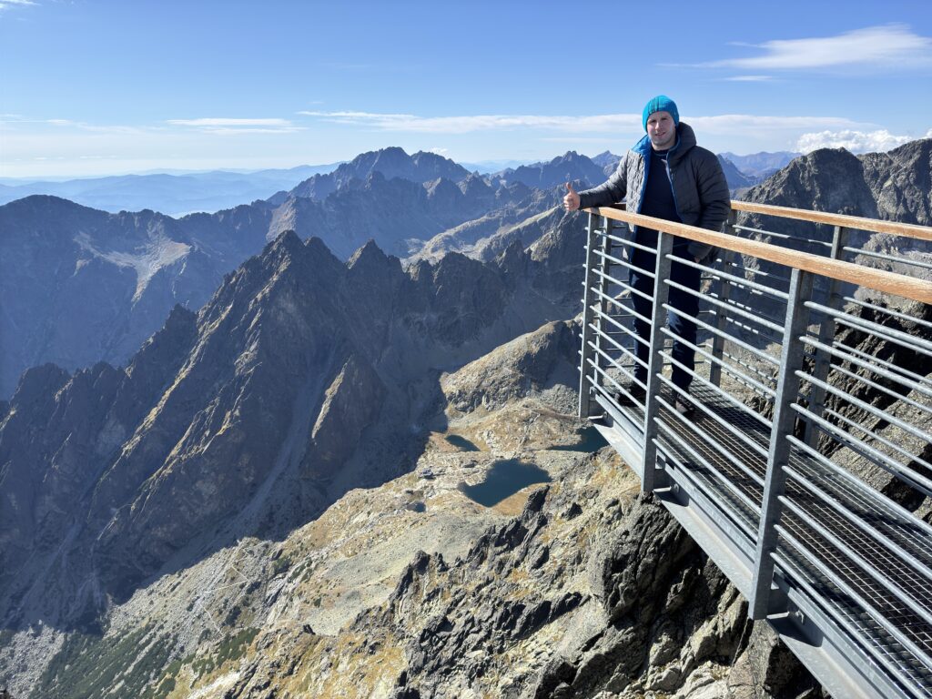 Man giving a thumbs up on a viewing platform at Lomnický štít, surrounded by dramatic High Tatras mountain peaks under a clear sky
