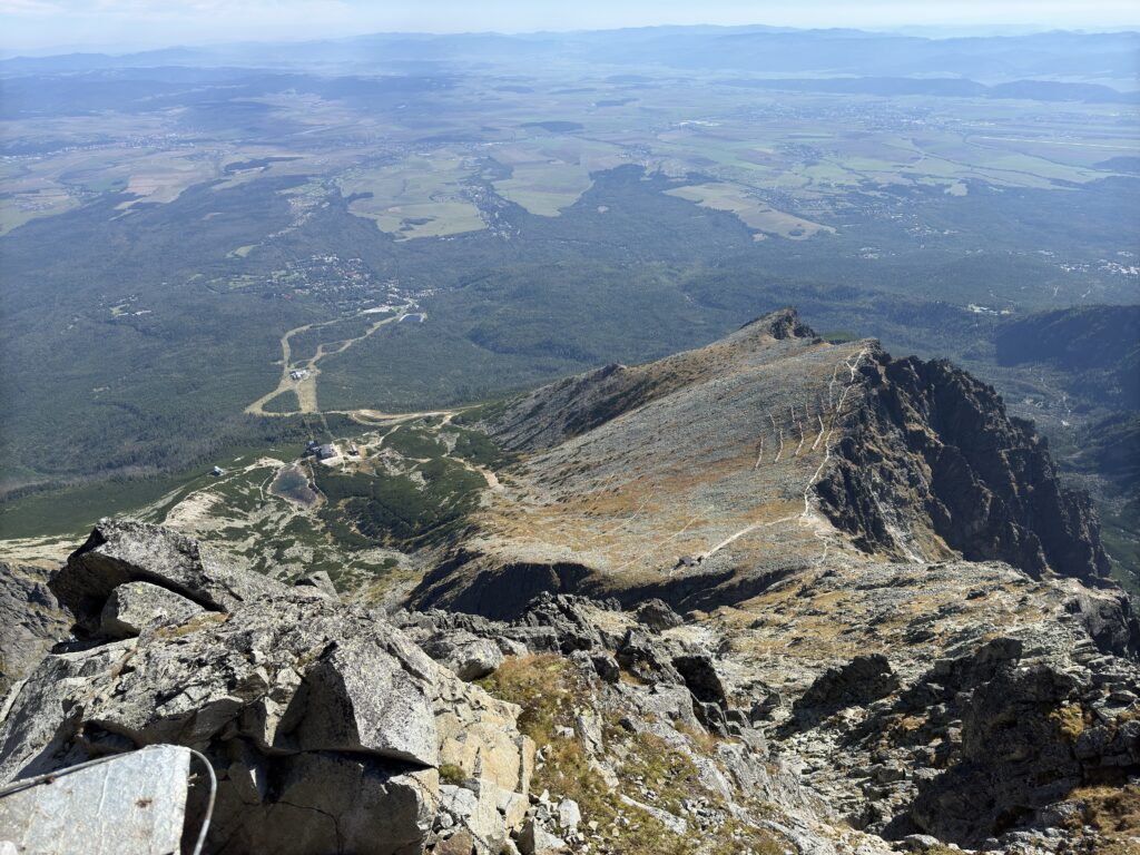 Panoramic view from a mountain peak overlooking a rocky ridge, winding trails, forests, and distant towns in the valley below