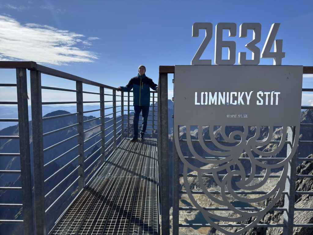 Man standing on the viewing platform at Lomnický štít, 2634 meters above sea level, with mountain peaks and a clear sky in the background