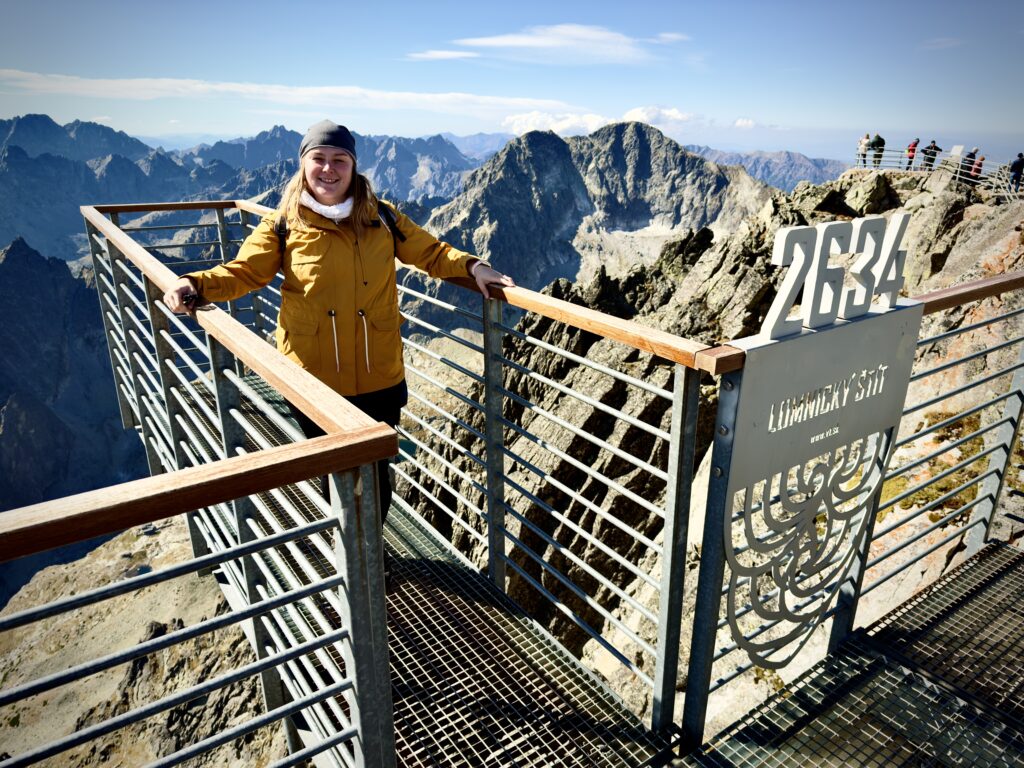 Woman in a yellow jacket standing on a viewing platform at Lomnický štít, 2634 meters above sea level, with dramatic mountain peaks in the background