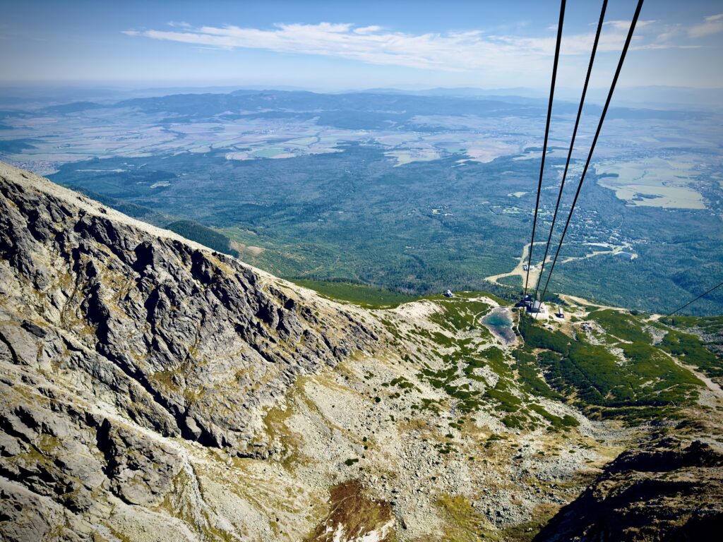Steep rocky mountain slope with cable car lines leading down to a valley with forests, fields, and distant towns