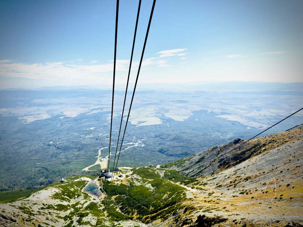 View from a mountain showing cable car lines descending toward a valley with forests, fields, and distant towns