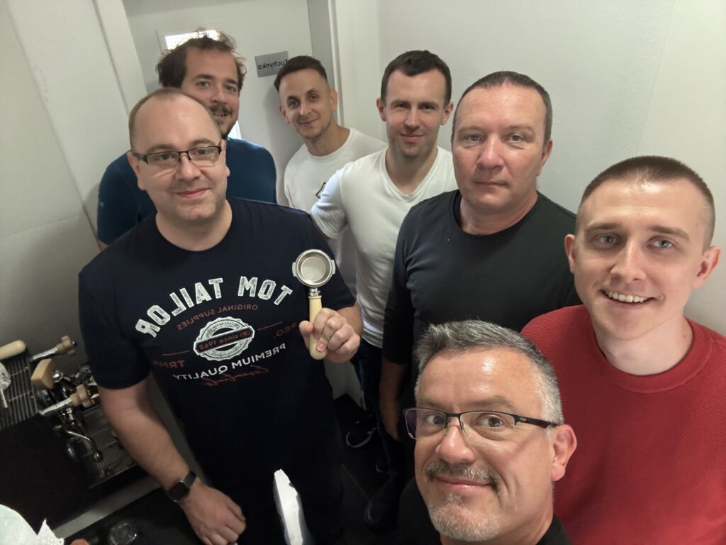 Group of seven smiling men standing in a small room, with one of them holding a portafilter near an espresso machine