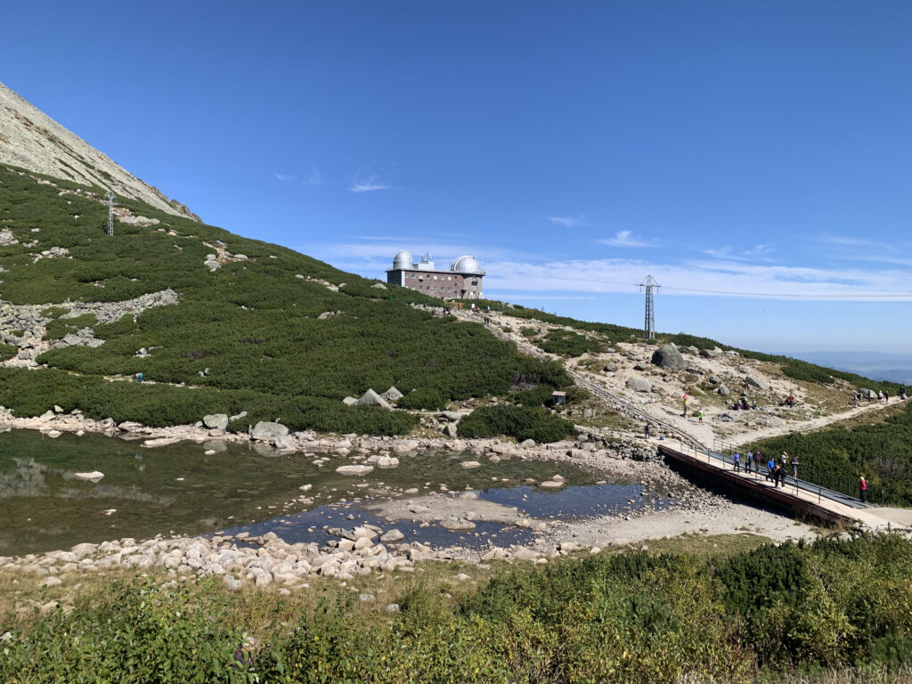 Mountain landscape with an observatory building on a hill, a small pond in the foreground, and hikers walking along a trail and bridge