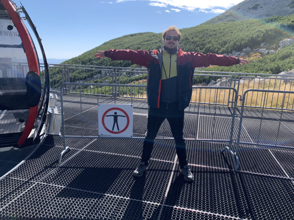 Man standing on a mountain platform with arms outstretched in front of a sign that prohibits this exact pose
