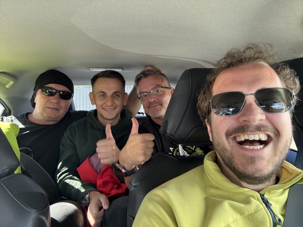 Four smiling men inside a car, three sitting in the backseat and one in the front passenger seat wearing a yellow jacket, with two showing thumbs up and all enjoying a cheerful moment