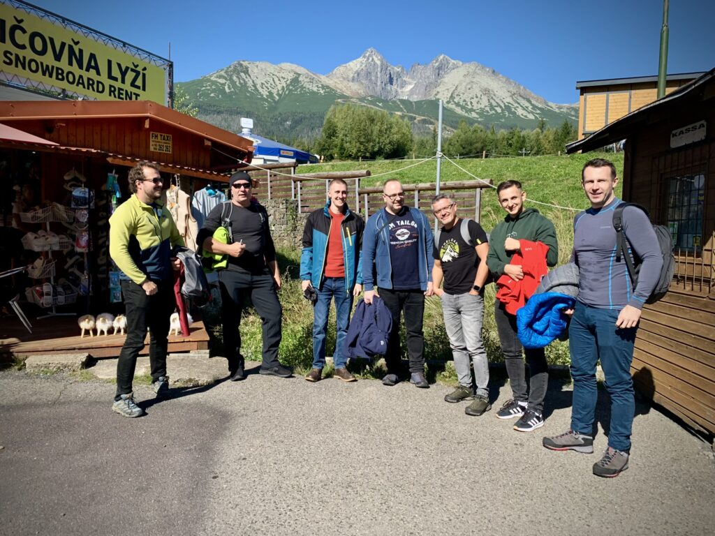 Group of men posing in front of a ski and snowboard rental shop with mountains in the background on a sunny day