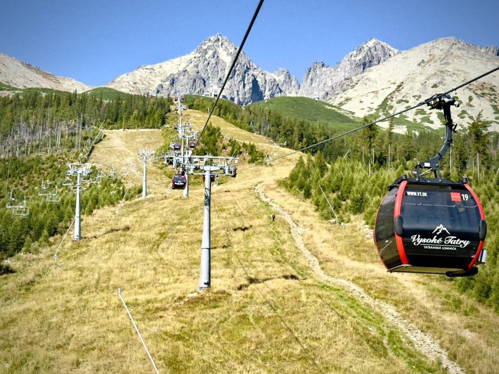 Red gondola lift with "Vysoké Tatry – Tatranská Lomnica" logo ascending a grassy mountain slope with high peaks in the background