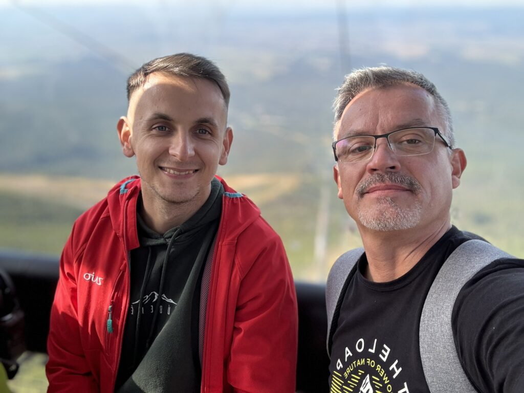 Two smiling men taking a selfie inside a cable car with a blurred mountainous landscape in the background