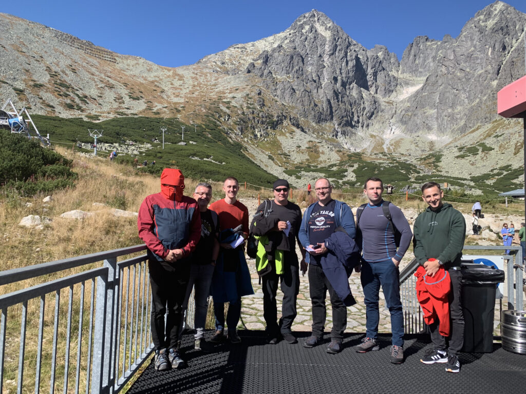 Group of men posing on a mountain platform with rocky peaks and cable car infrastructure in the background on a sunny day