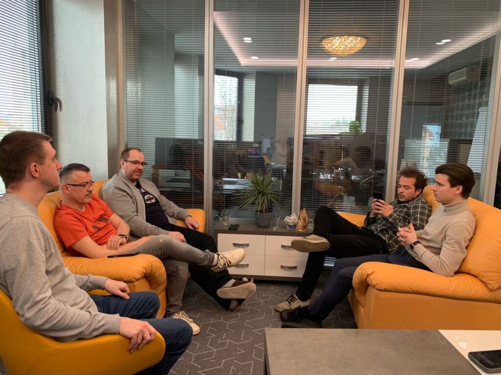 Five men are sitting on orange sofas in a modern office lounge, engaged in casual conversation. One is using a phone while others listen or talk. A glass wall with blinds separates the lounge from a meeting room in the background.