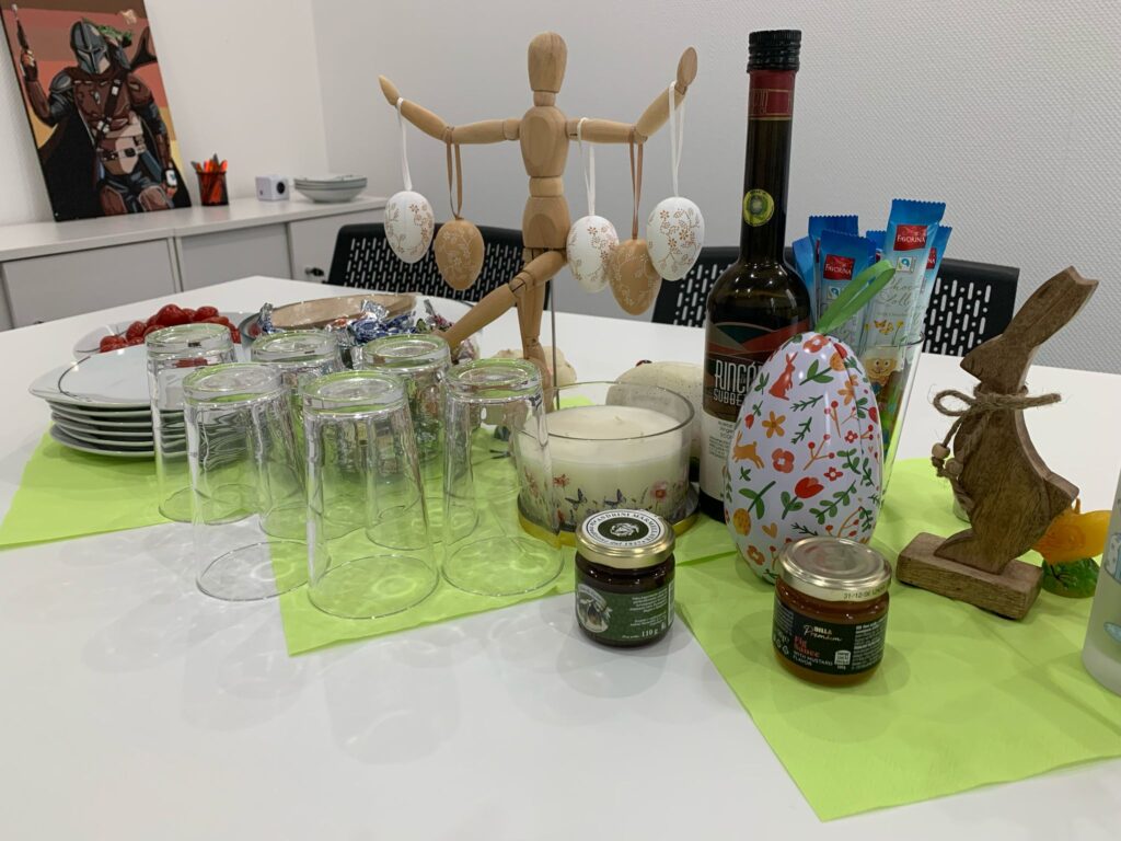 A festive Easter table with glasses, plates, candy, decorated eggs, a wooden bunny, and snacks. A wooden figure holds hanging Easter eggs. A colorful egg and treats are arranged on green napkins.
