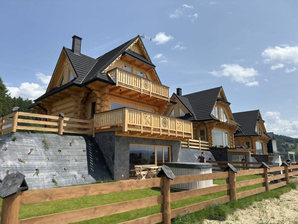 A large wooden cabin with balconies and black rooftops in the Polish mountains, rented for Evolit's teambuilding. The cabin is surrounded by a wooden fence and grassy area under a clear blue sky.