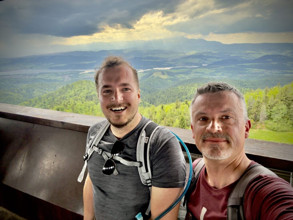 Two Evolit team members smiling for a selfie at a scenic viewpoint during a hike in the Polish mountains. Behind them is a vast landscape of green hills, a lake in the distance, and dramatic clouds breaking sunlight through the sky.