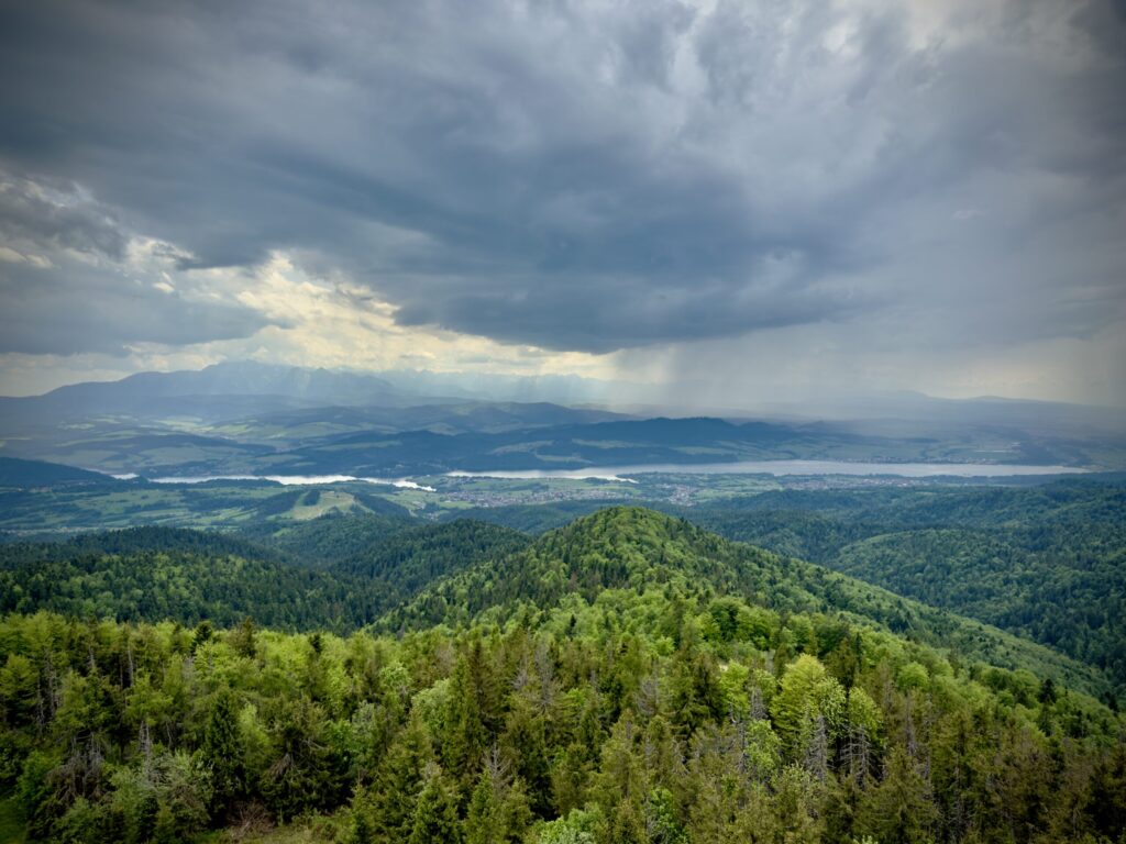 Panoramic view of lush green hills and forests in the Polish countryside, with a distant lake and low-lying villages beneath a dramatic cloudy sky. Sunlight breaks through in places, illuminating parts of the landscape.
