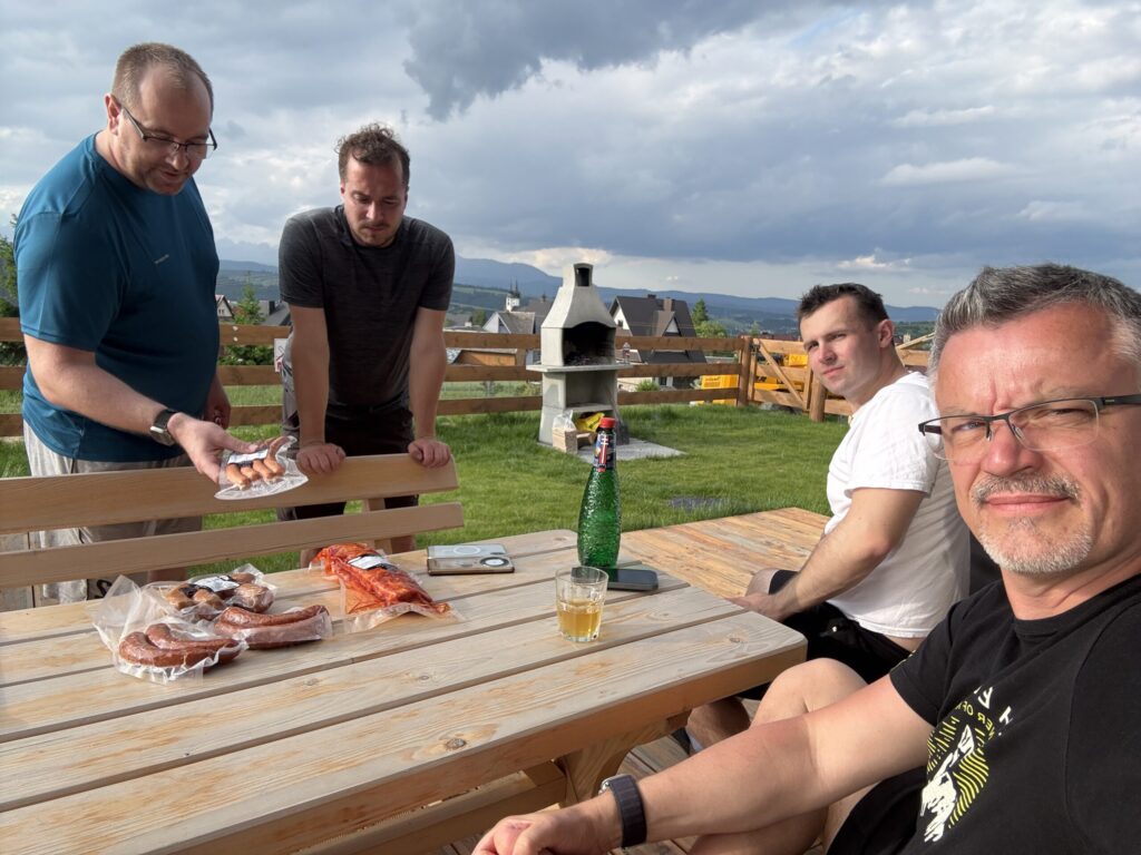 Four men preparing for a barbecue outside a wooden cabin in Poland. Two are sitting at a wooden table, and two are standing while checking meat and sausages. A grill stands in the background with mountains and village rooftops visible under a partly cloudy sky.