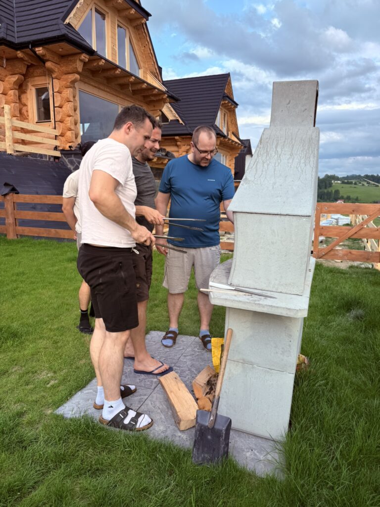 Four men gathered around a concrete grill in front of a wooden cabin, grilling food on skewers during a teambuilding event in Poland. Firewood and a shovel lie nearby on the grass.