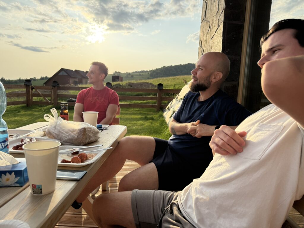 Three men relax on a wooden terrace during sunset, enjoying food and drinks after a barbecue. A rural landscape and soft sunlight create a peaceful atmosphere.