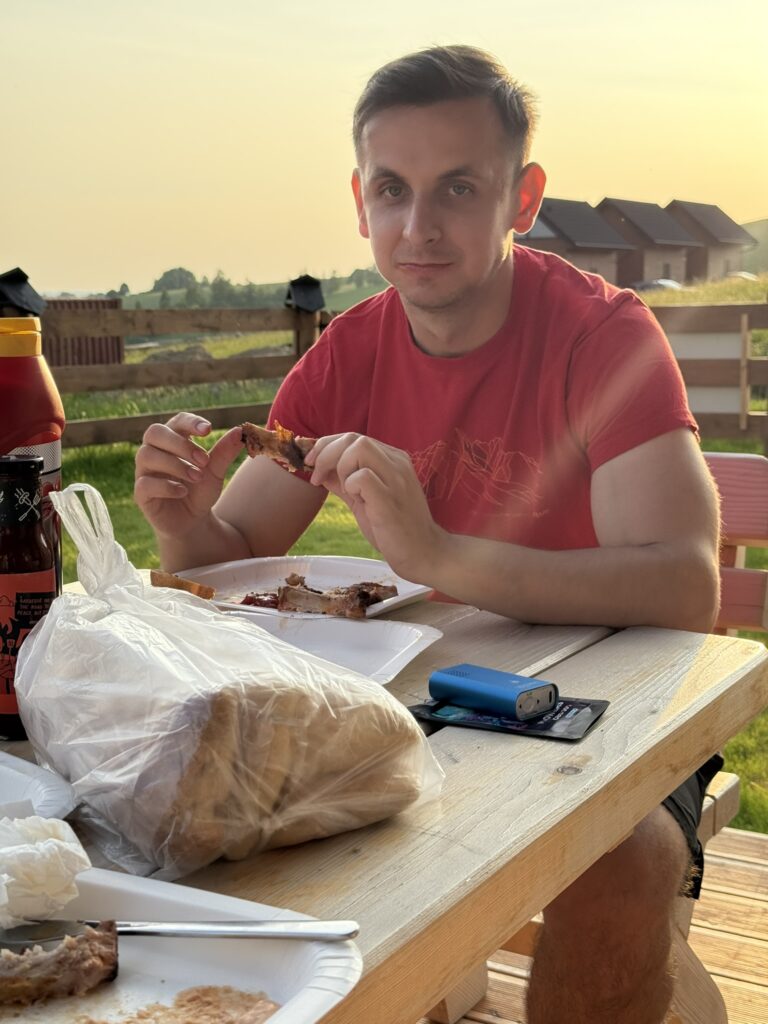 Man in a red T-shirt sitting at a wooden table outdoors, eating grilled meat during sunset, with a loaf of bread, sauce, and disposable plates in front of him.