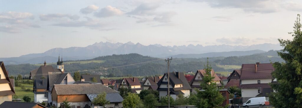 Panoramic view of a village in southern Poland with houses and church spires in the foreground, and the Tatra Mountains visible in the distance under a partly cloudy evening sky.